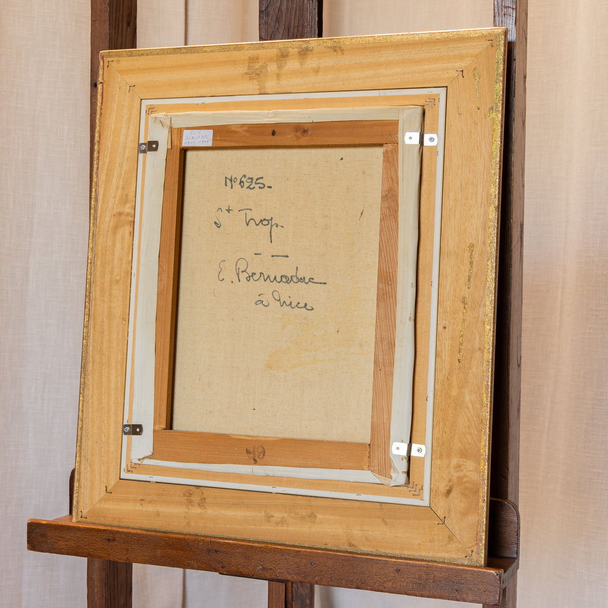 The back of a framed artwork on a wooden easel, showing a light wood frame, cream inner matting, canvas backing, and handwritten inscription.
