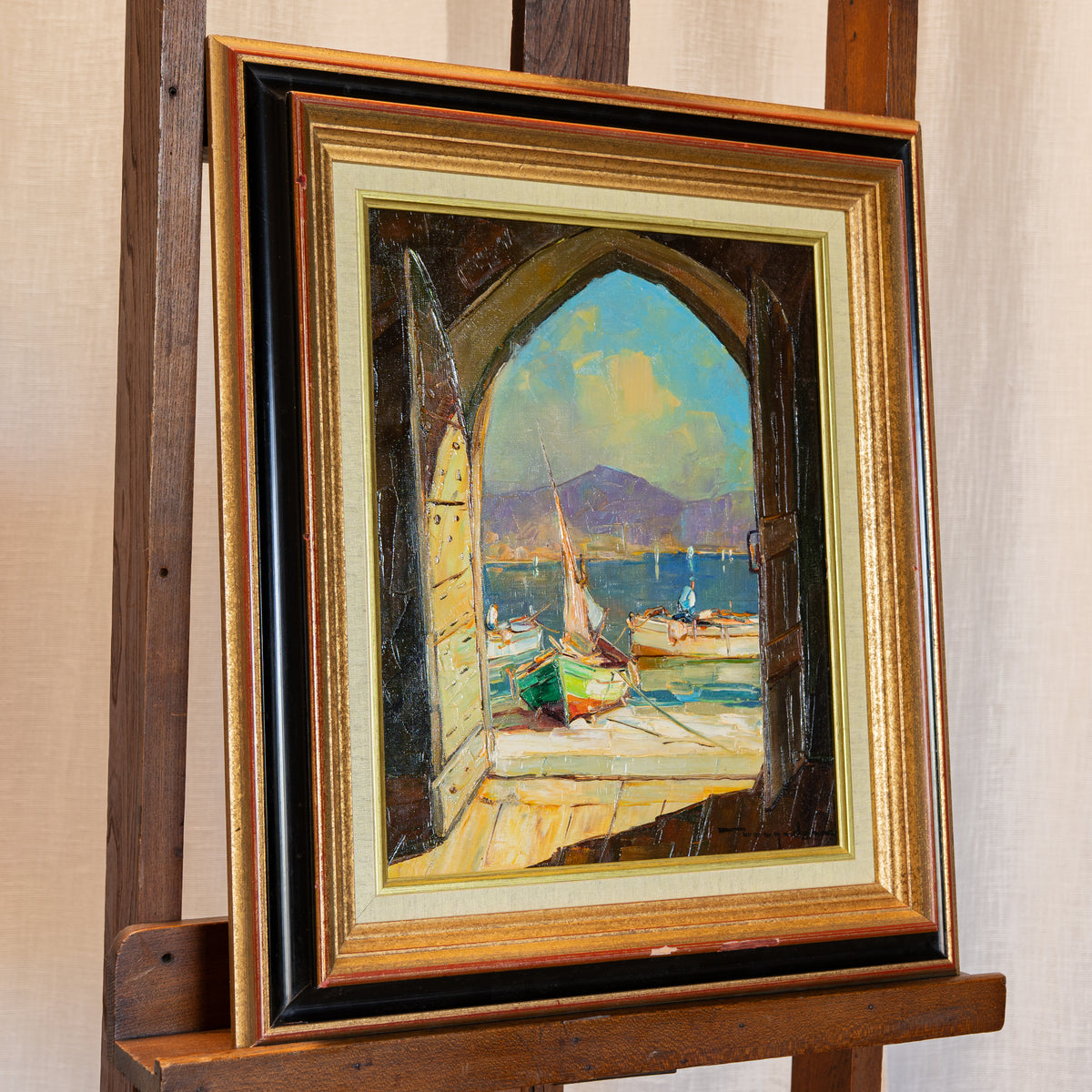 A framed oil painting depicting a scenic harbor view seen through an open wooden archway, displayed on a wooden easel. The painting features colorful boats on the water under a blue sky, and the frame is ornate with gold, black, and cream tones.