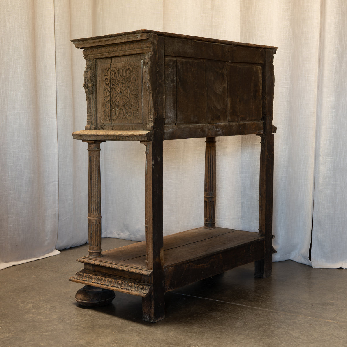 An antique dark wood two-tier console table or credenza with ornate carved details on the upper cabinet front and fluted legs, photographed against a draped white fabric backdrop.
