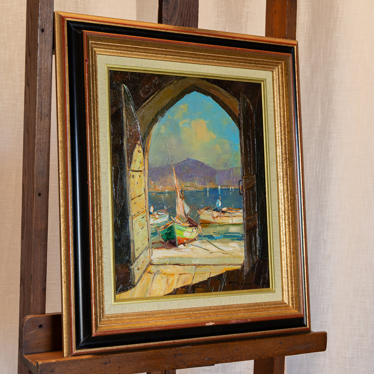 A framed oil painting of a harbor scene with boats and mountains, displayed on a wooden easel against a neutral fabric backdrop.