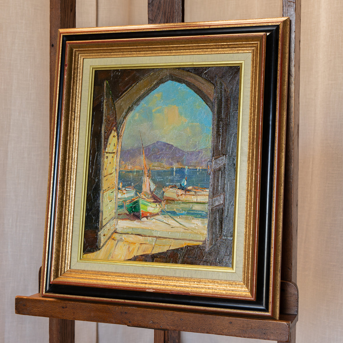 A framed oil painting depicting a harbor scene viewed through an arched doorway, displayed on a wooden easel. The painting features colorful boats and a blue sky, enclosed in a multi-layered gold, black, and beige frame.