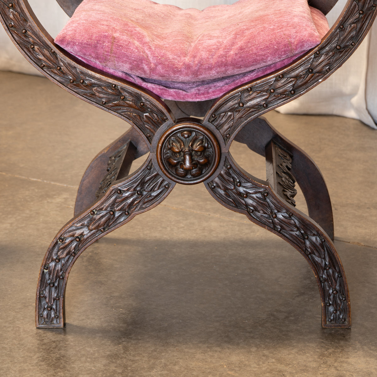 Close-up of an ornate carved wooden bench or stool with a pink velvet seat cushion, shown indoors on a concrete floor.
