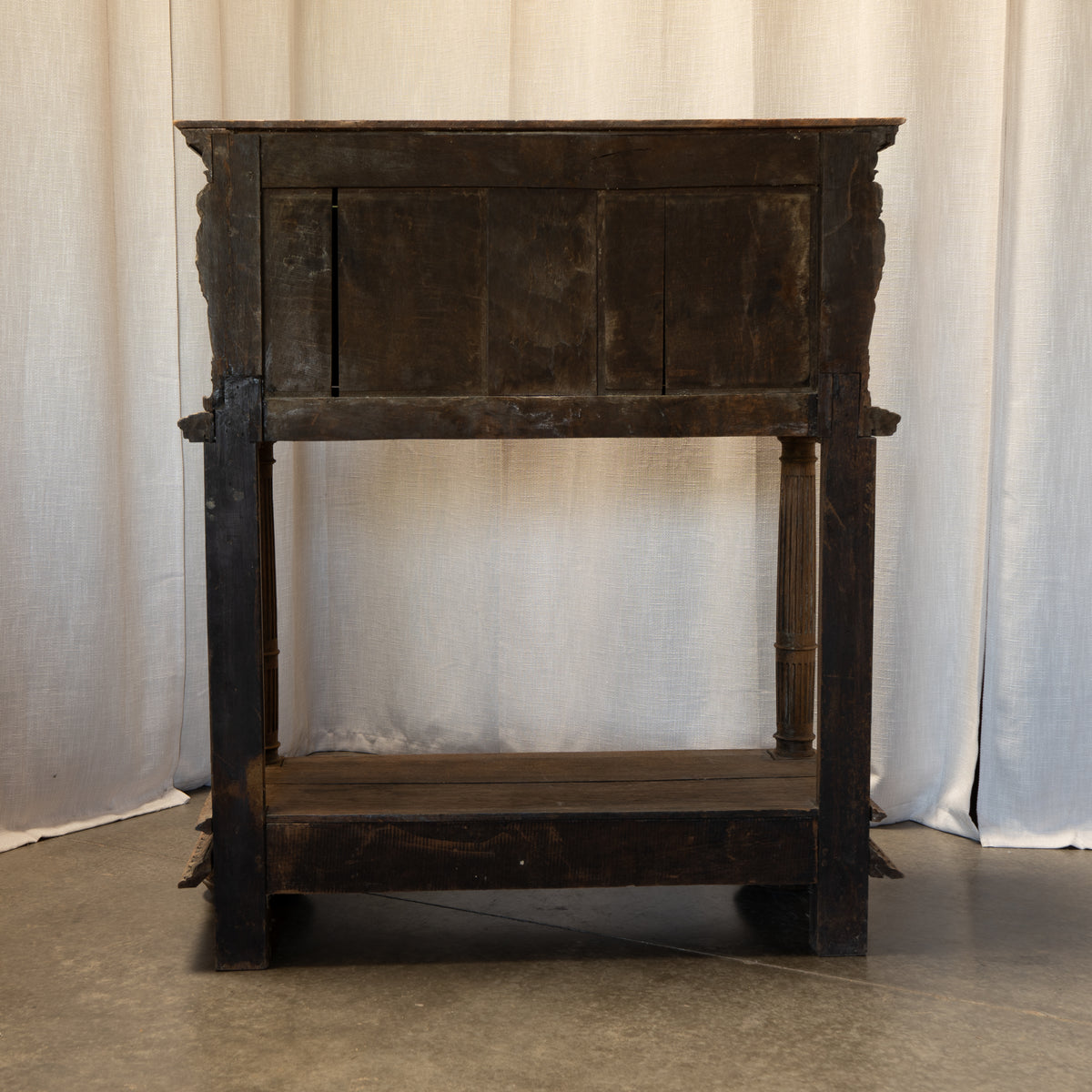 A dark rustic wooden side table or cabinet with a single upper door panel, open middle shelf, and carved legs, photographed against a draped off-white fabric backdrop.