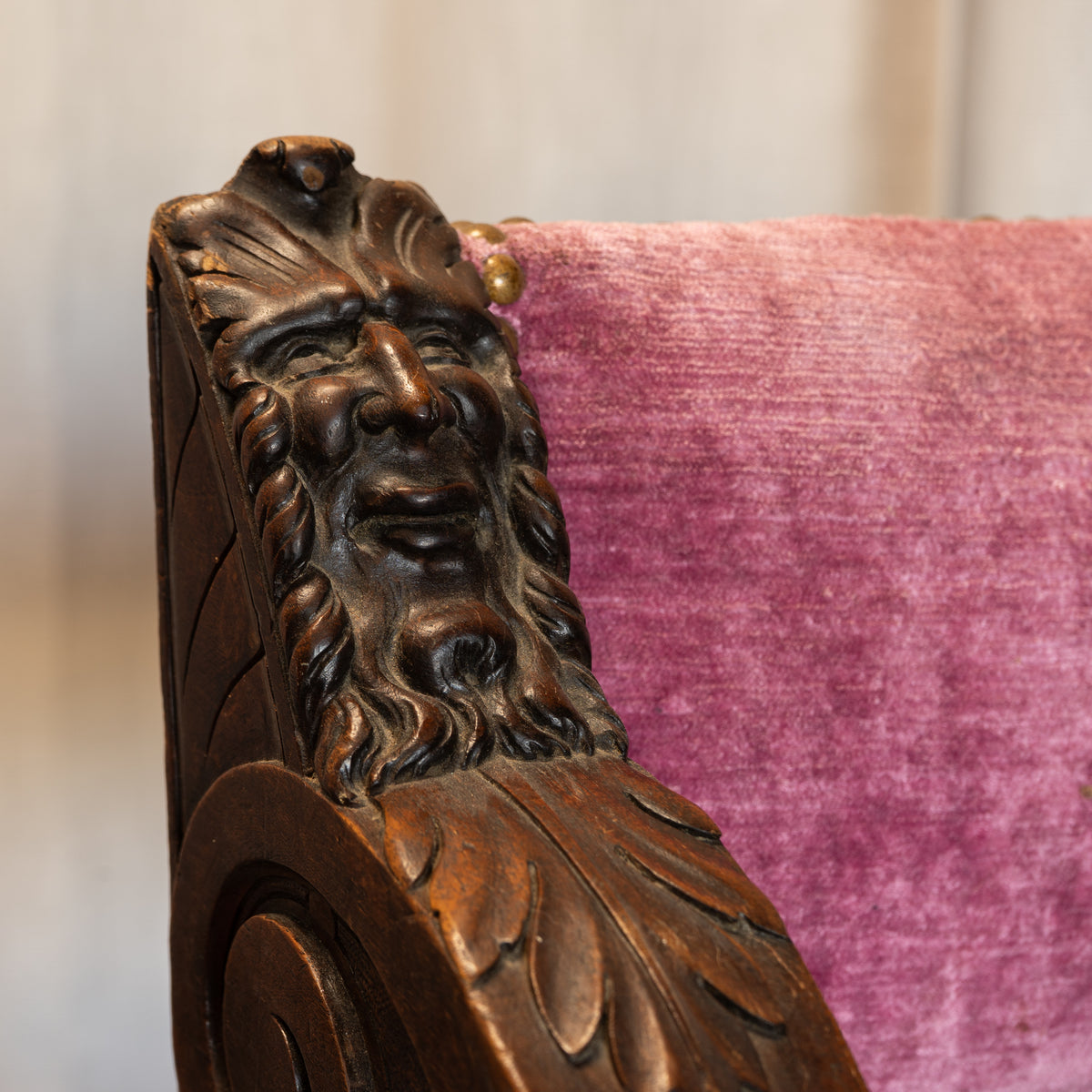 Close-up of an ornate carved wooden chair arm featuring a detailed face and leaf-like scrollwork, paired with a textured magenta-pink upholstered backrest.