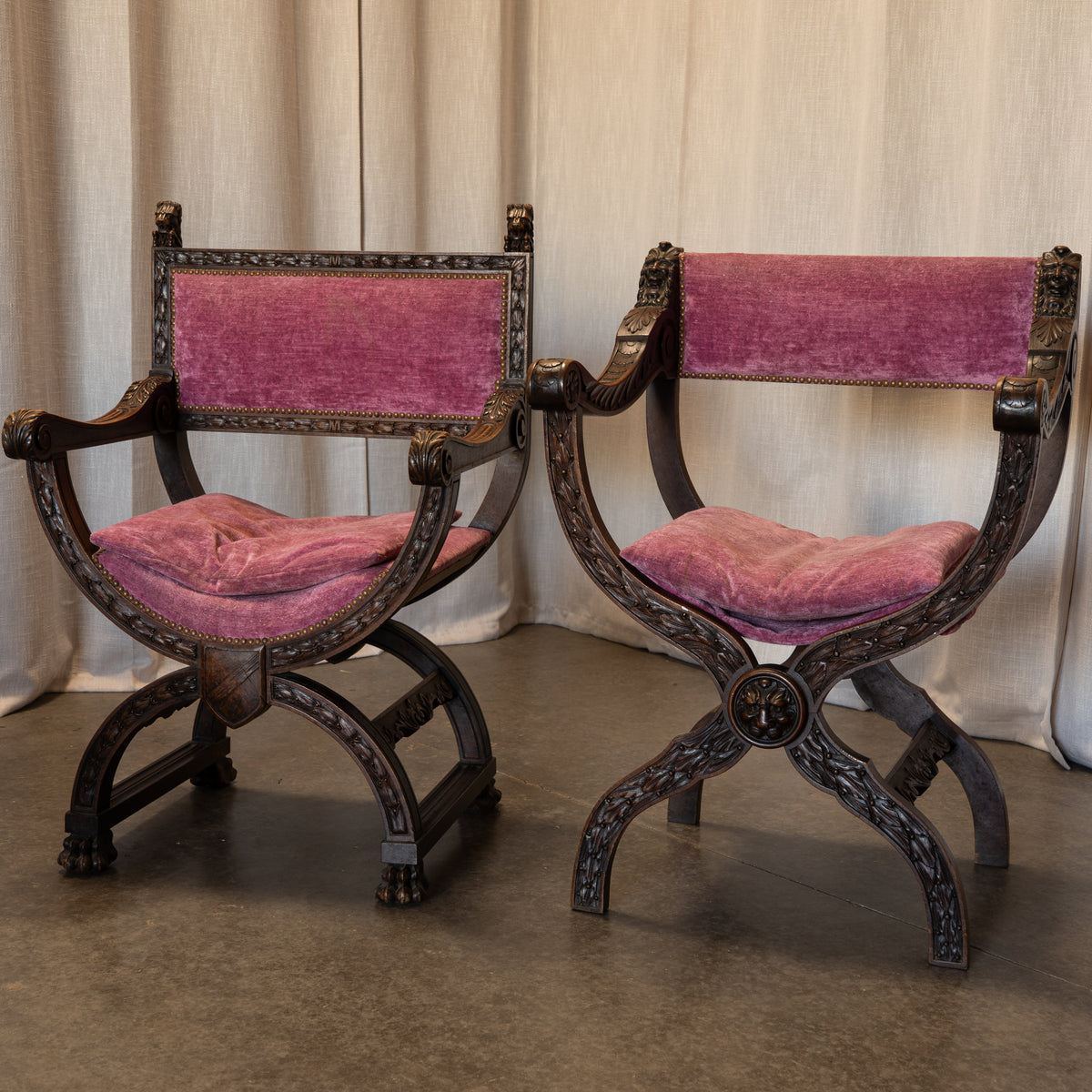Two ornate vintage-style wooden armchairs with carved dark wood frames and plush magenta velvet cushions, set against a neutral draped fabric backdrop.