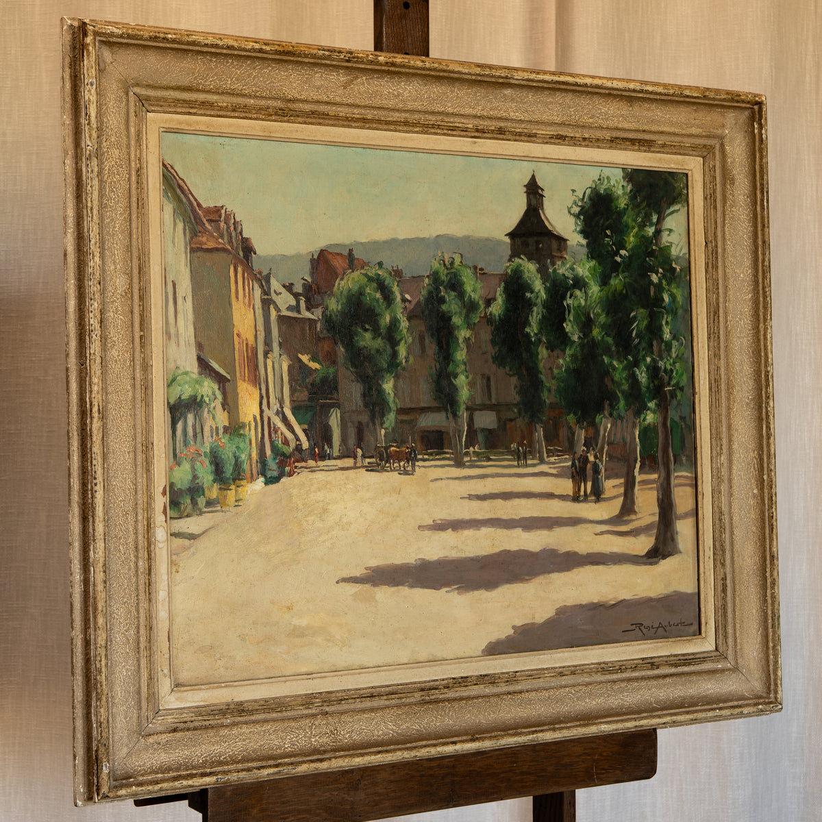 An oil painting of a European street scene with trees, buildings, and horse-drawn carriages, housed in an ornate distressed gold frame and displayed on a wooden easel.