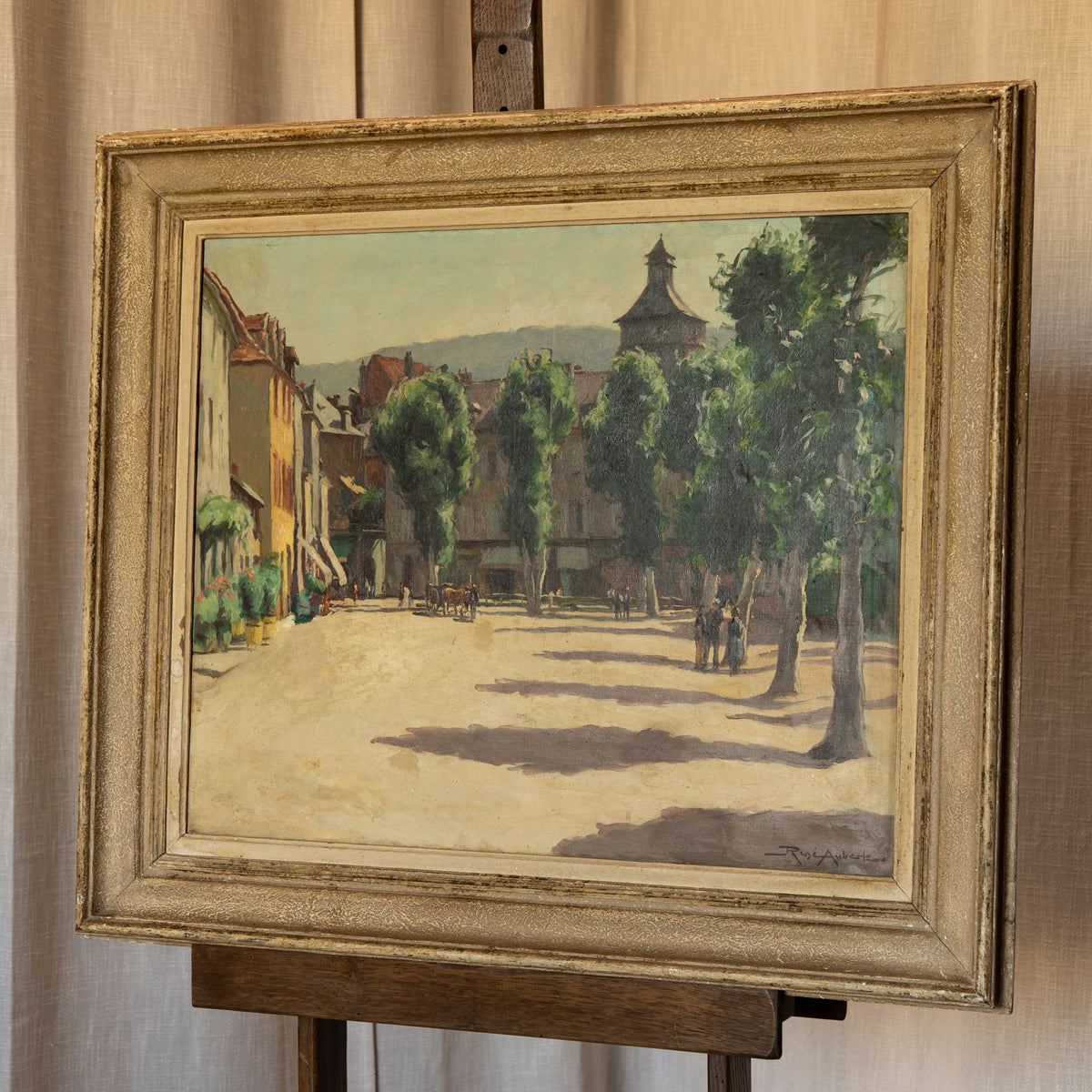 An oil painting of a European town square with trees, buildings, and horse-drawn carriages, housed in an ornate gold-toned frame and displayed on an easel.