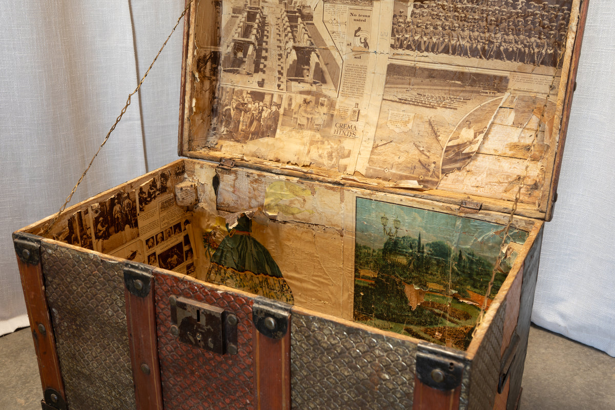 An antique wooden trunk with a diamond-plate patterned front and a hinged lid, shown open. The inside of the lid and sides are decorated with vintage newspaper clippings and a small landscape print.