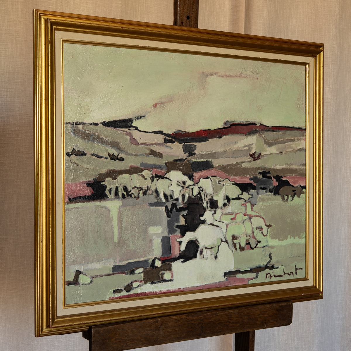 A framed painting depicting a herd of white sheep in a stylized landscape with muted green, beige, and pink tones, housed in a wide gold frame and displayed on a wooden easel against a beige curtain background.