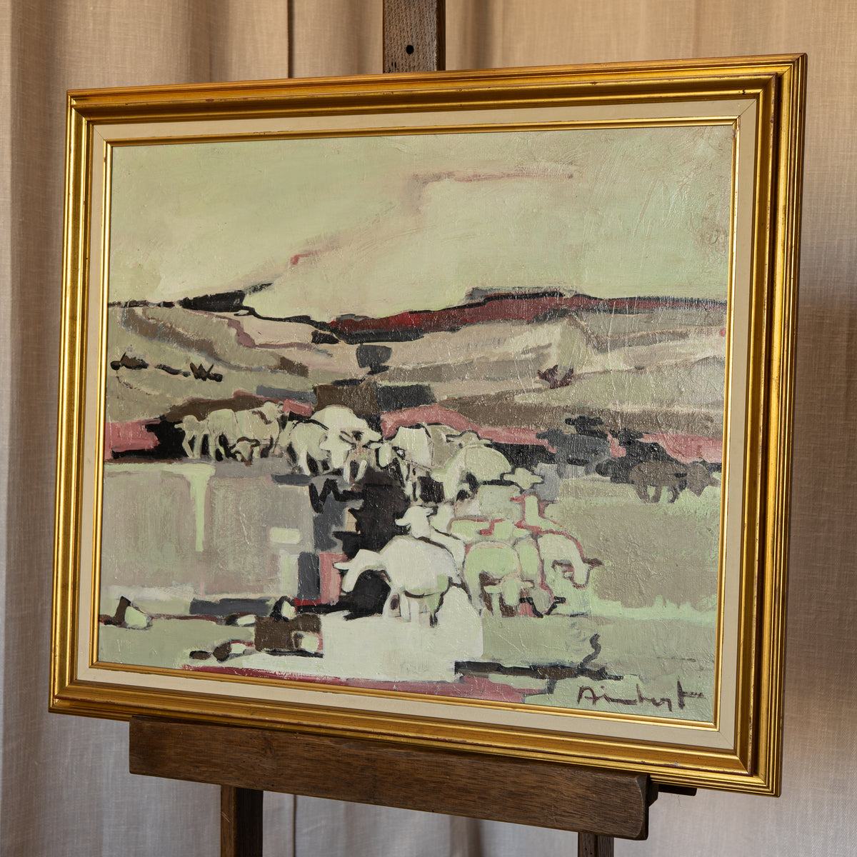 A framed landscape painting depicting a herd of white sheep on a hillside, housed in a wide gold frame and displayed on a wooden easel against a beige curtain background.
