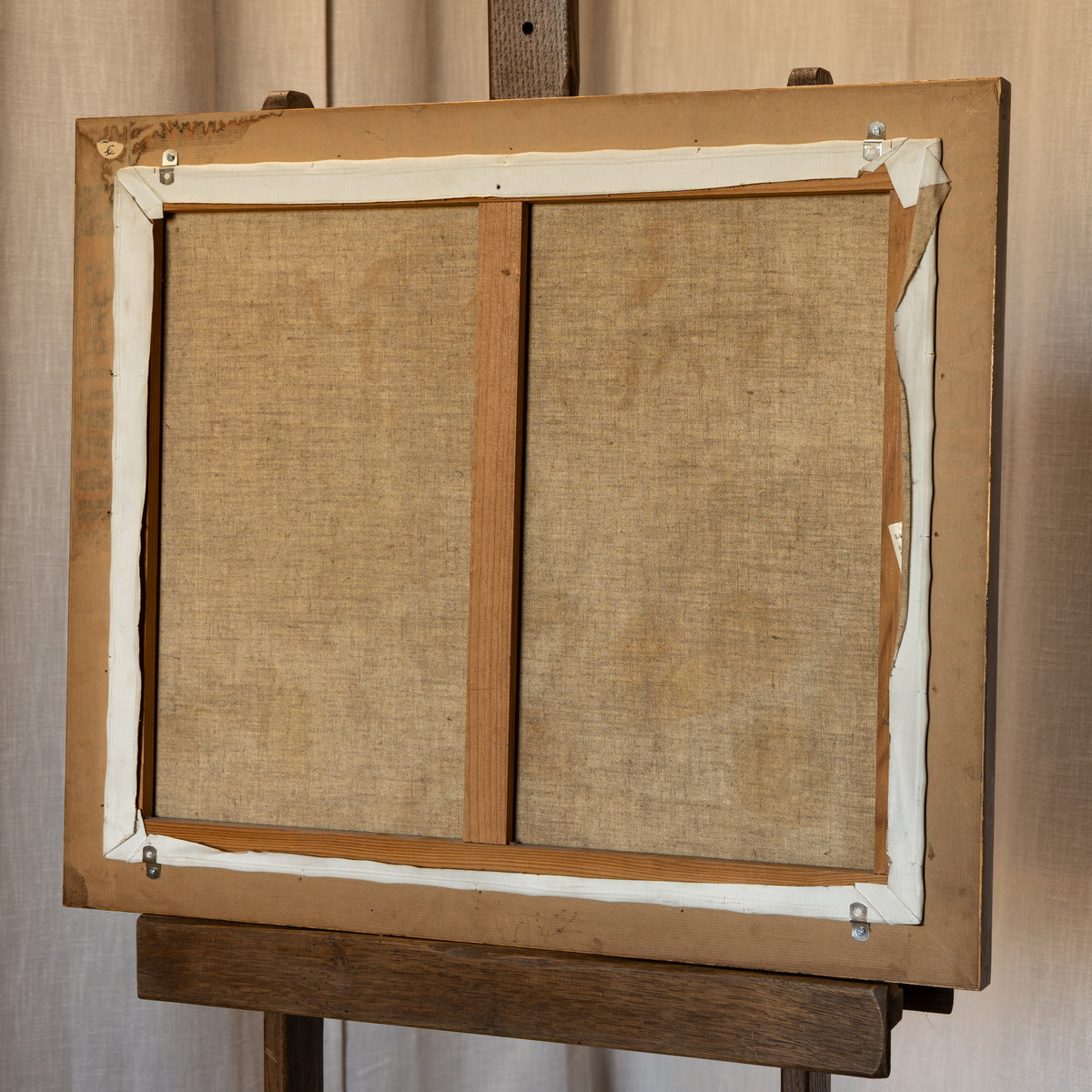 Back view of a framed canvas or print on an easel, showing the tan linen fabric stretched over a wooden frame with white tape along the border.