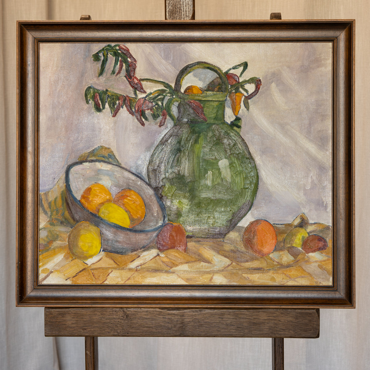 A framed still life oil painting on an easel, depicting a green vase with dried leaves and oranges, a bowl of oranges, and other fruits on a patterned tablecloth.