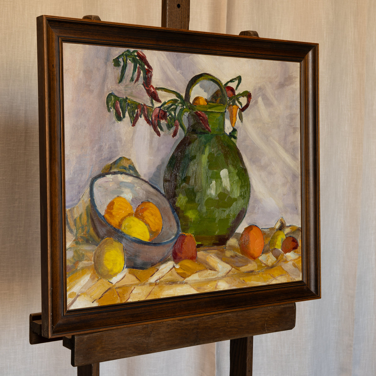 A framed still life oil painting on an easel, featuring a green pitcher, a bowl of lemons and oranges, and a vase with dried flowers against a light background.
