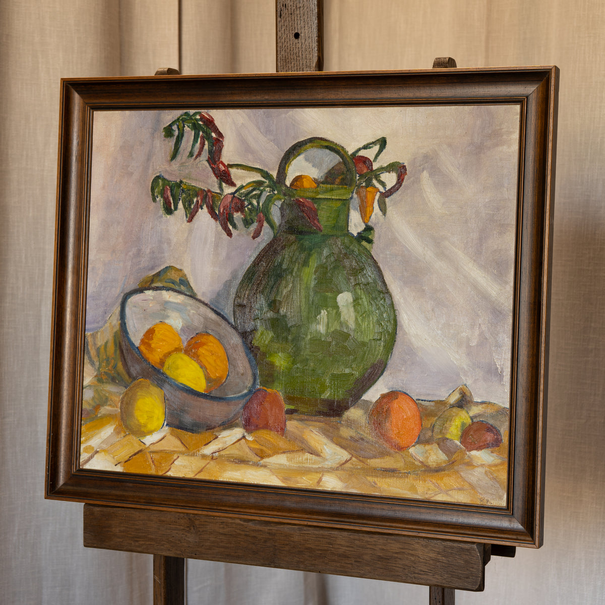 A framed still life oil painting on an easel, featuring a green pitcher, a bowl of oranges, and fruit on a draped table against a light background.