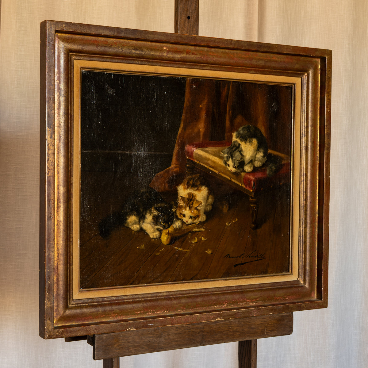 A framed vintage oil painting depicting two kittens on a wooden floor, mounted on a wooden easel against a neutral beige wall.