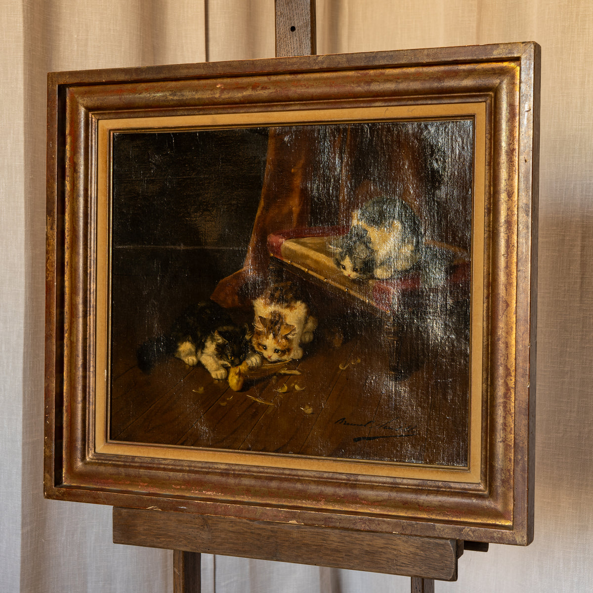 A framed oil painting depicting two cats on a wooden floor in a dimly lit interior, mounted on a wooden easel against a light curtain background.