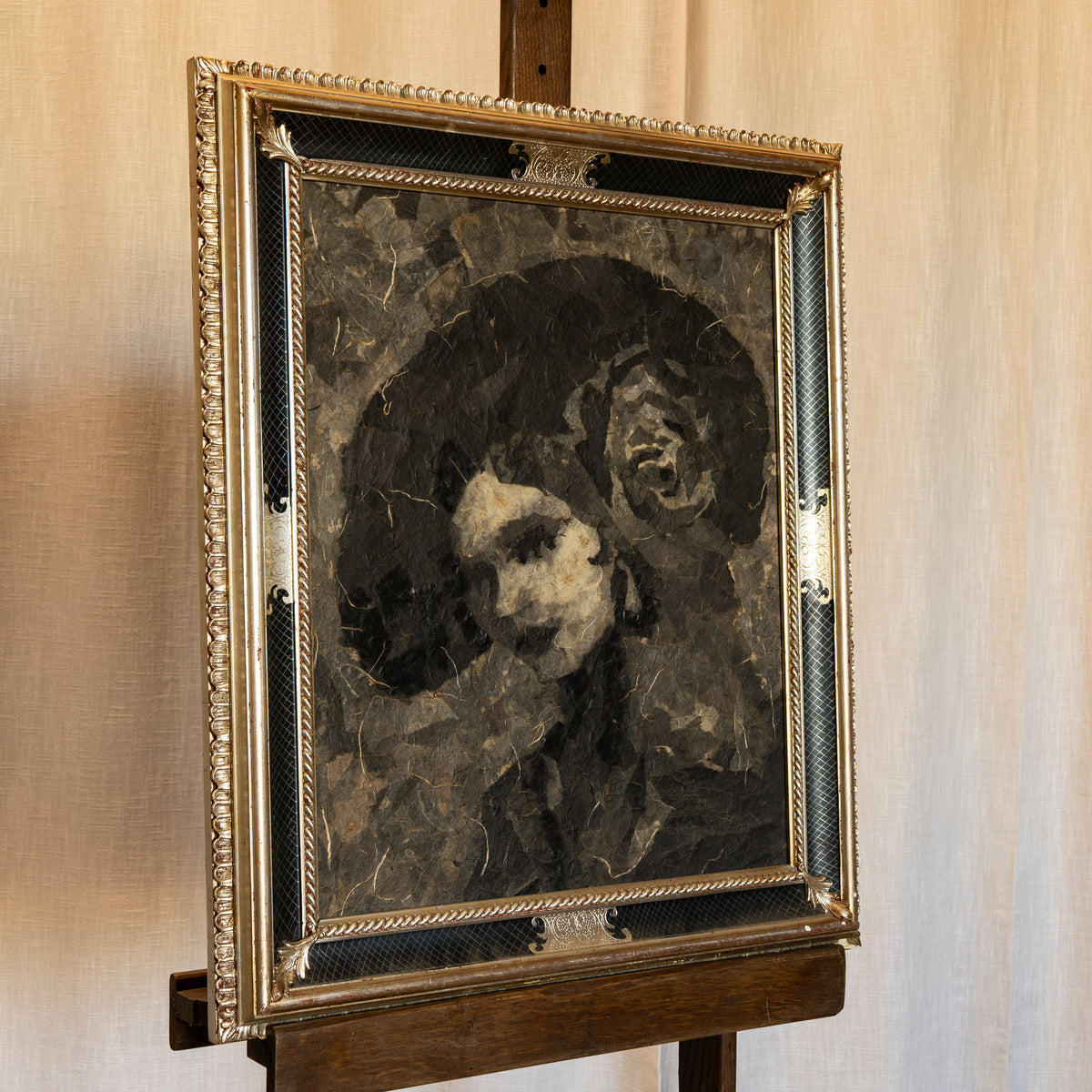 A framed portrait painting of a woman with dark hair and a white dog, displayed on a wooden easel against a beige fabric backdrop.