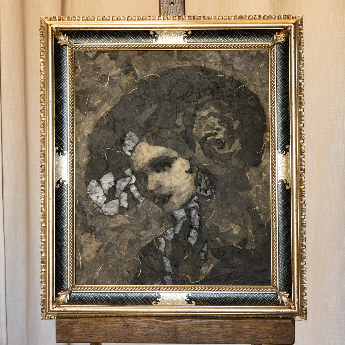 A framed portrait painting of a woman with a man in the background, rendered in dark earthy tones with white accents, set in an ornate gold and black frame on a wooden stand against a beige wall.