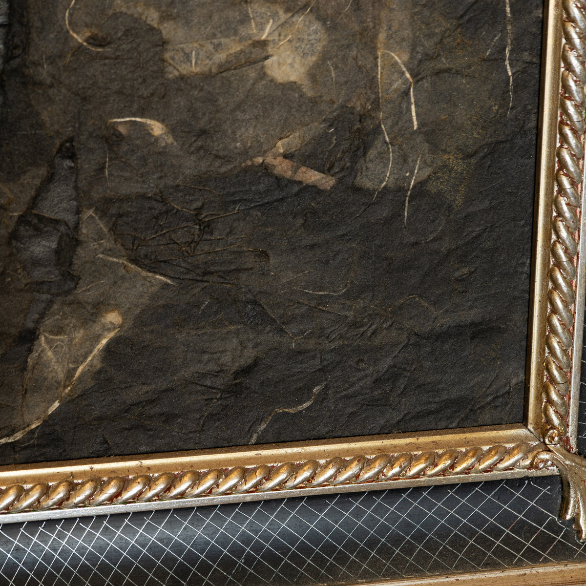 Close-up of a framed artwork showing a dark textured surface with a decorative gold rope-style frame corner and a small portion of a patterned surface below.