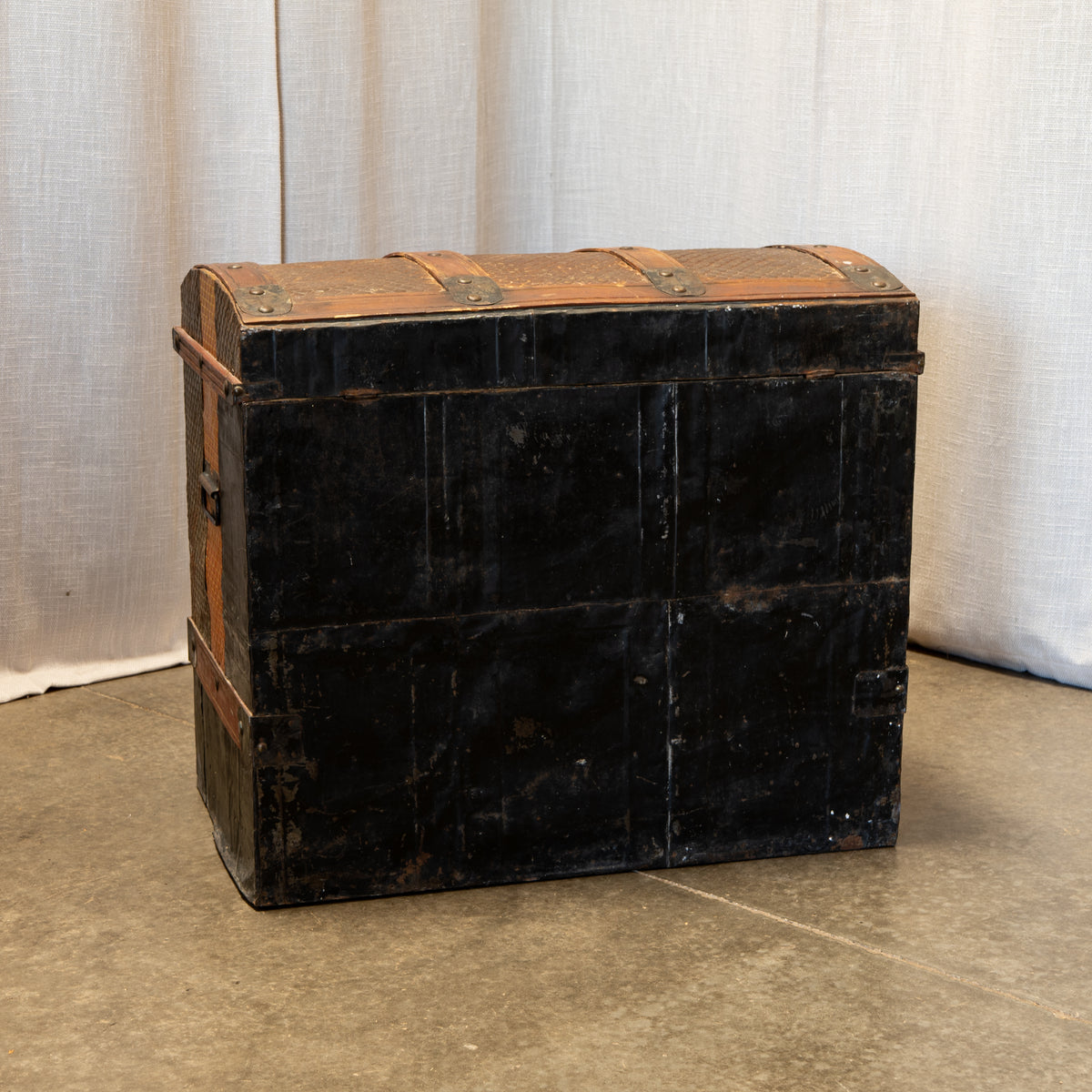 A vintage black wooden trunk with weathered brown leather straps and metal corner reinforcements, set against a neutral draped fabric backdrop.