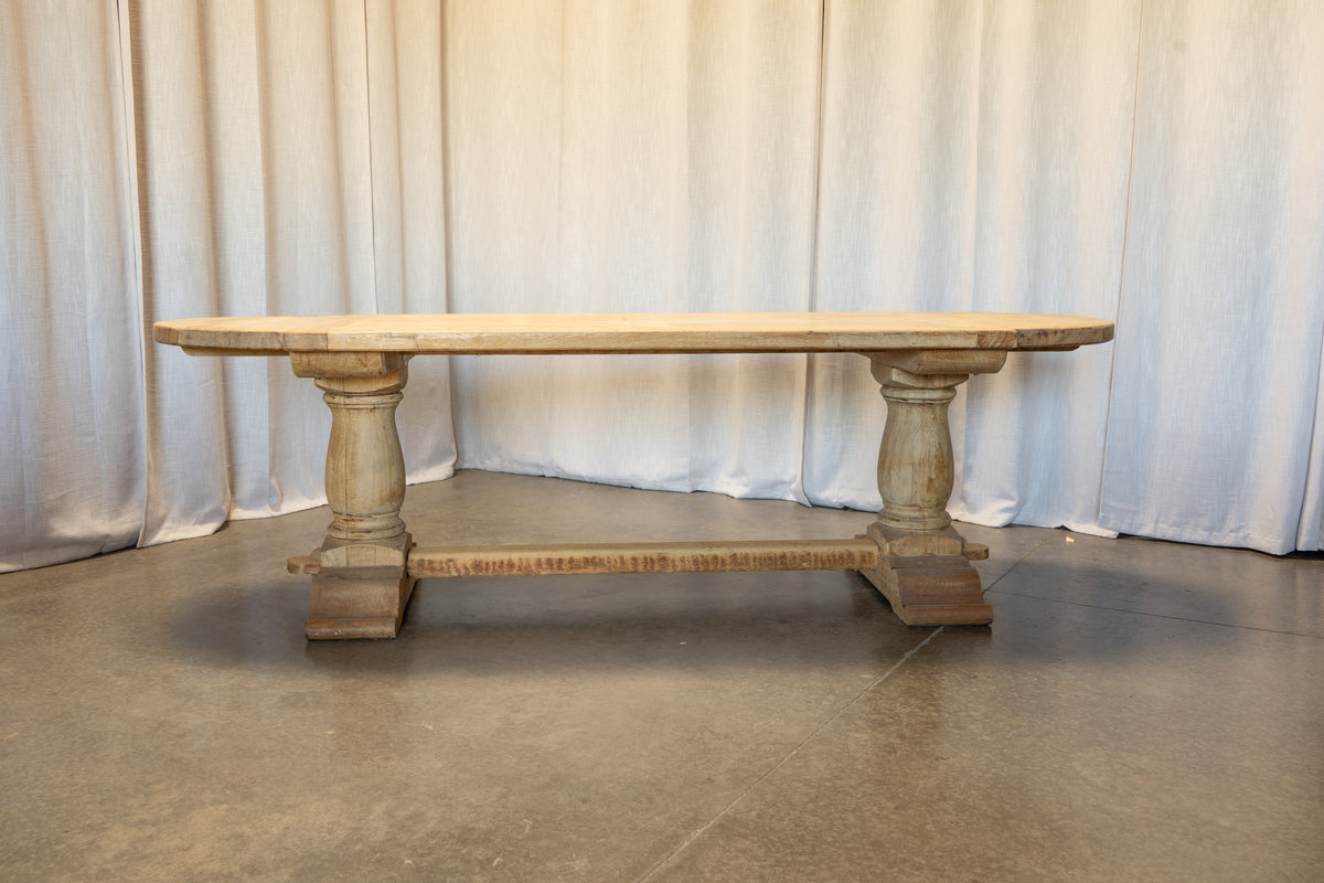 A rustic wooden dining table with a light natural wood top and turned pedestal legs, set against a draped white fabric backdrop on a concrete floor.