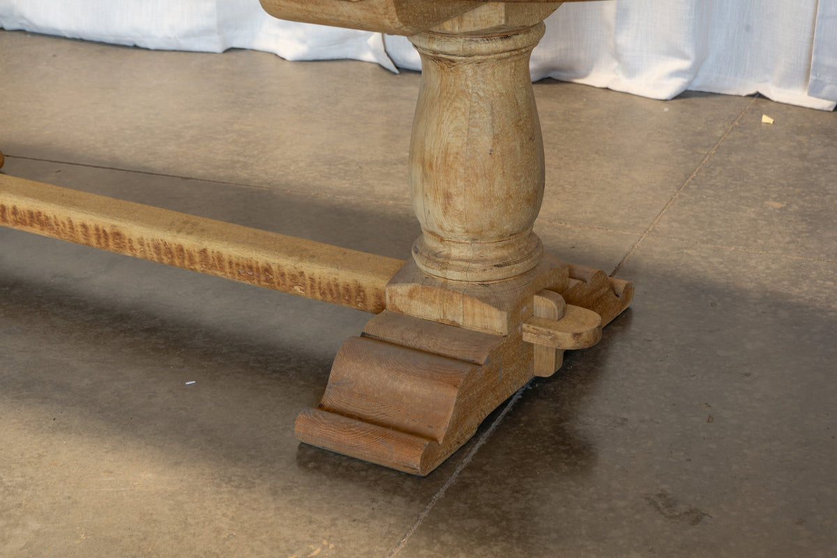 Close-up of a rustic wooden table leg with a turned pedestal base and carved feet, resting on a concrete floor.