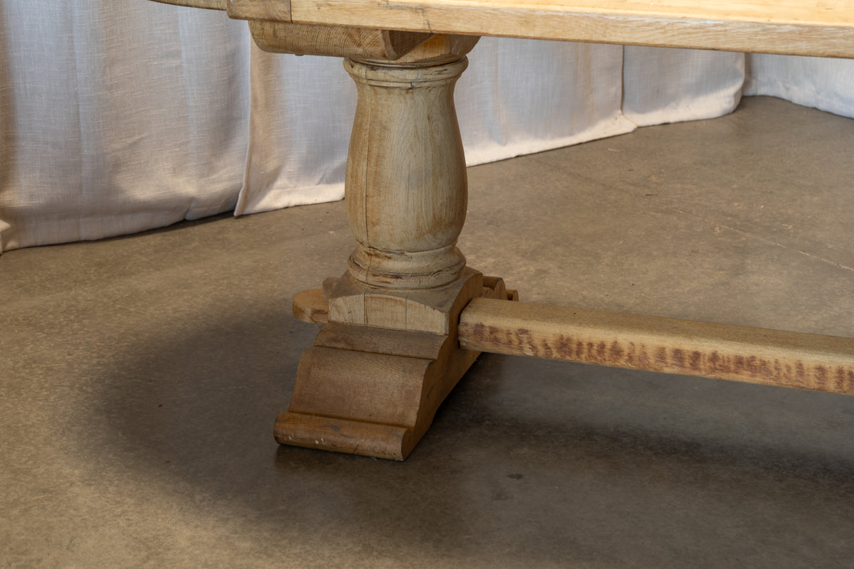 Close-up of a rustic light wood dining table leg and stretchers on a concrete floor with draped fabric in the background.