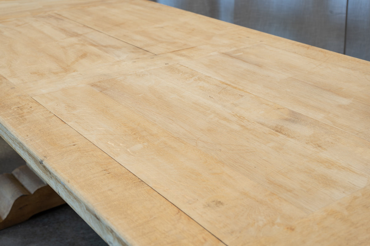 Close-up of a light natural wood tabletop with visible grain and panel seams, photographed indoors with part of a chair leg and a gray wall in the background.