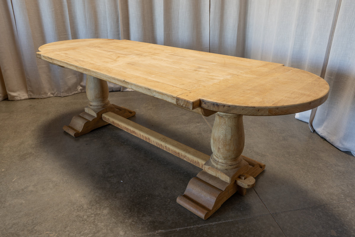 An oval wooden dining table with a light natural wood top and carved trestle-style legs, photographed indoors against draped fabric and a concrete floor.