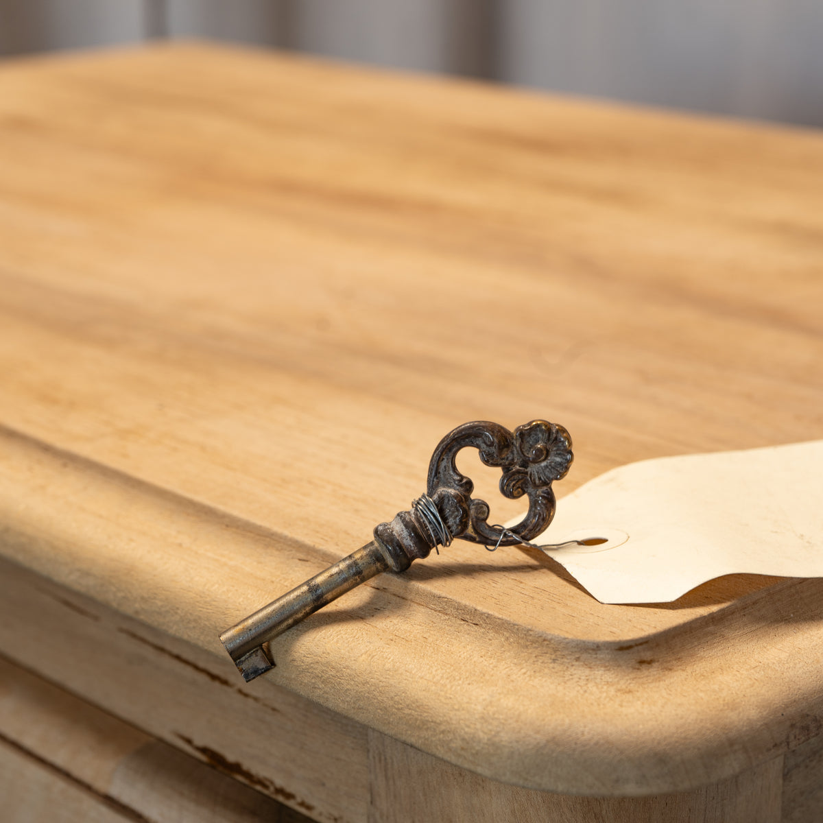 An ornate antique-style metal key with a dark patina rests on the corner of a light wood table next to a small beige paper tag.