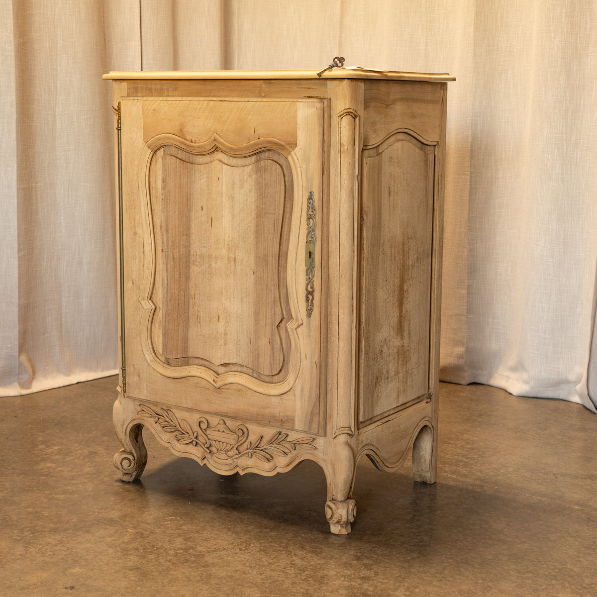 A distressed vintage French-style wooden cabinet with carved details and curved legs, finished in a light beige wood tone with a light wood top, set against a neutral draped fabric backdrop.