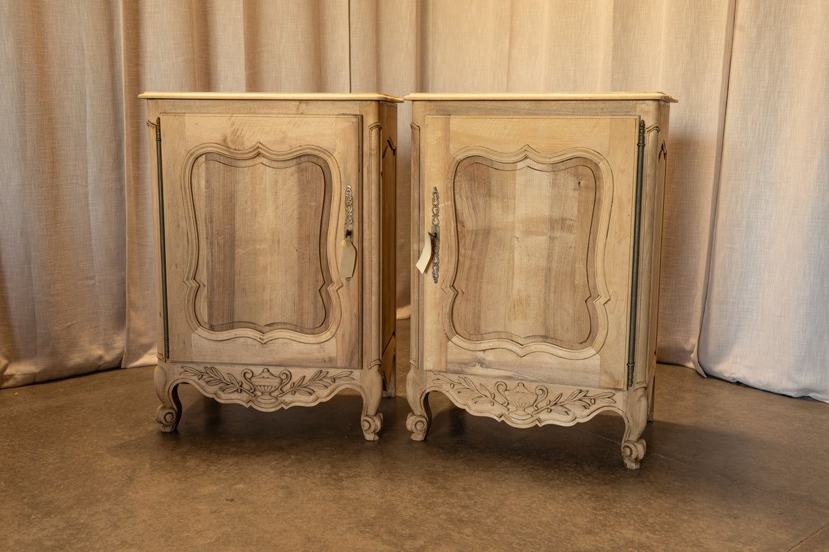 A pair of vintage French-style nightstands with carved details and a distressed beige finish, set against a neutral draped fabric backdrop.