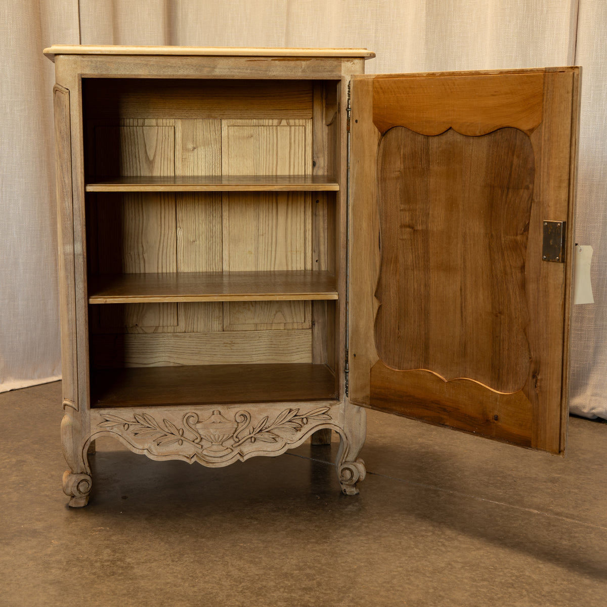 A vintage French-style cabinet with a distressed off-white carved frame and a warm brown wooden door, shown with the door open to reveal three interior shelves.
