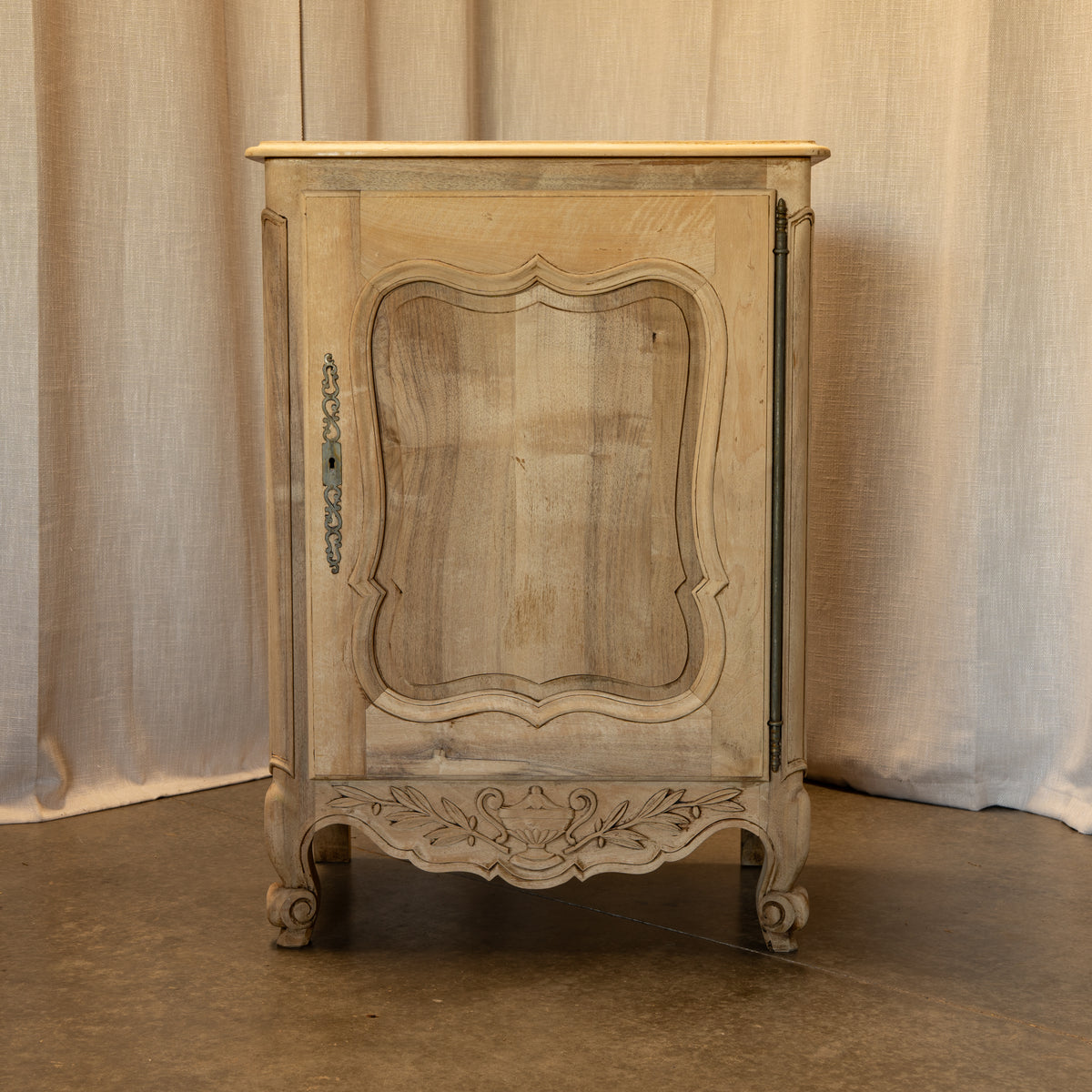 A distressed light beige wooden cabinet with ornate carved details, a curved top, and a single glass-paneled door, set against a neutral draped fabric backdrop.