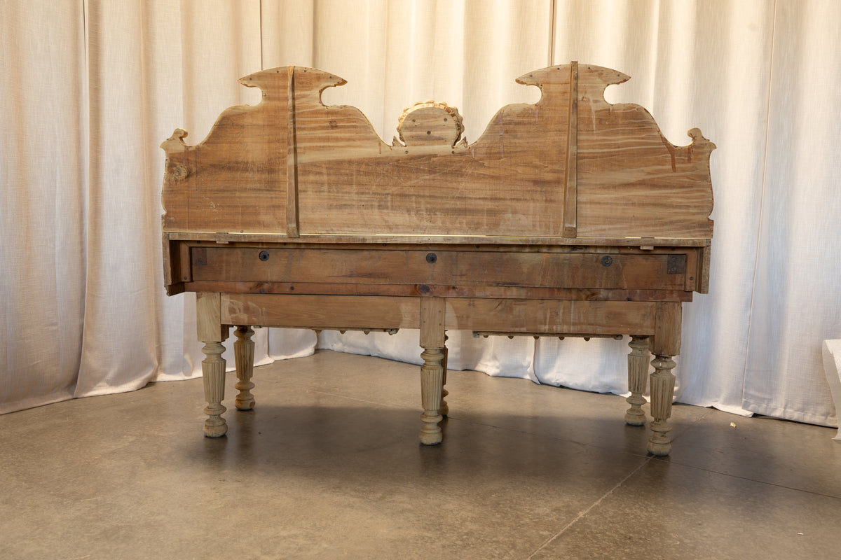 A rustic wooden bench with a tall carved headboard-style backrest, turned legs, and a weathered finish, set against a white curtain backdrop.