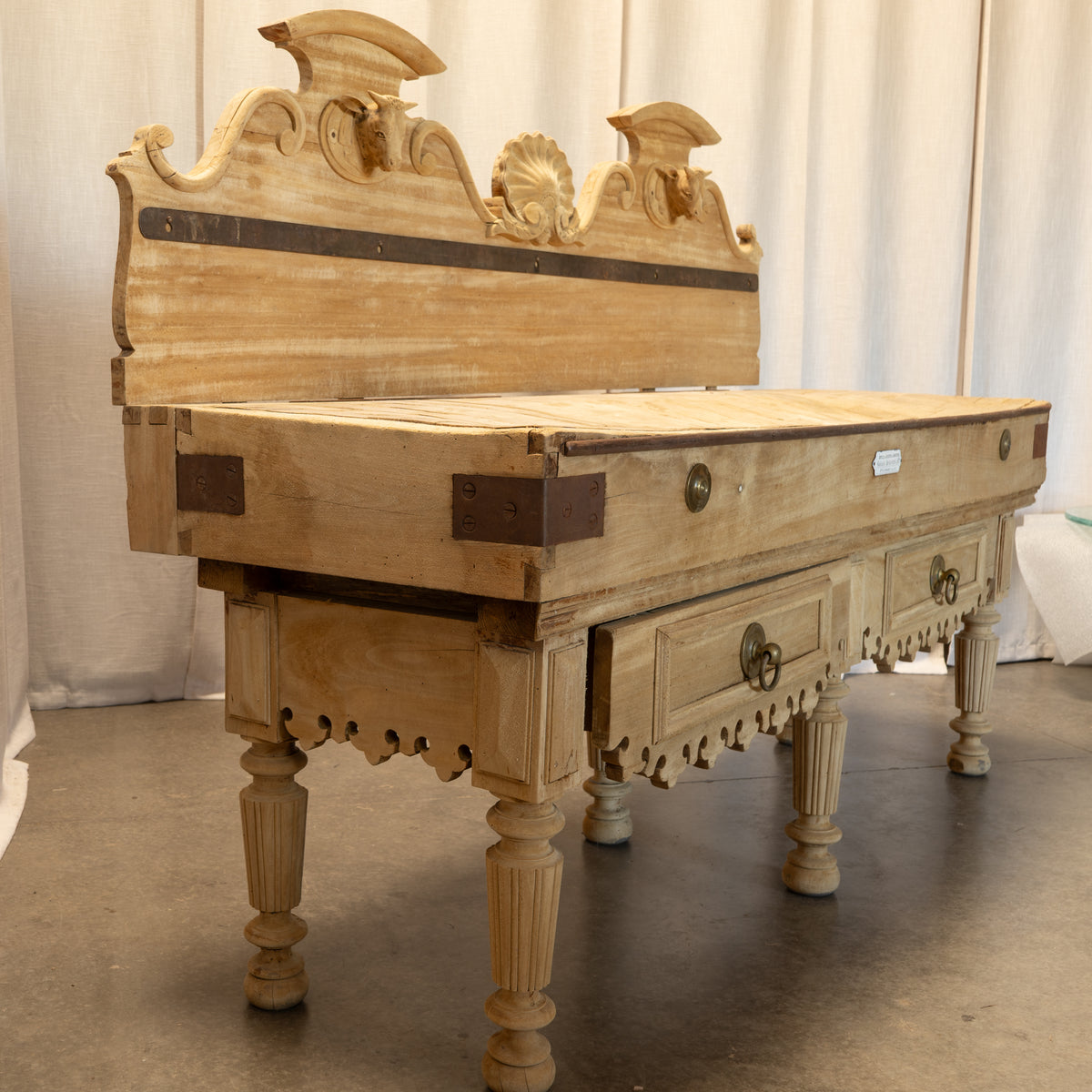 An ornate carved wooden chest of drawers with a high decorative headboard, featuring light natural wood, dark metal brackets, and turned legs, photographed indoors against a draped curtain backdrop.