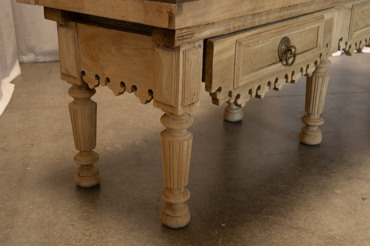 A close-up of a light natural wood table, showing carved legs, a drawer with a brass pull, and decorative scalloped apron details, set on a concrete floor.