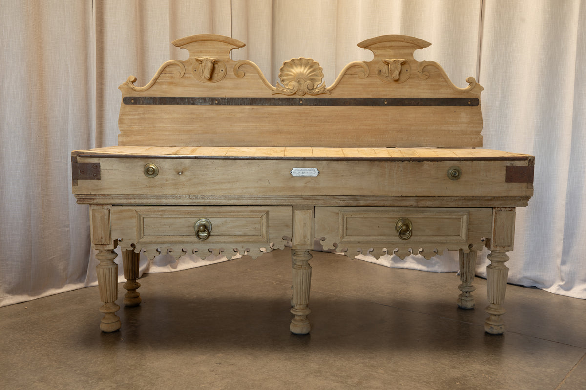 An antique carved wooden writing desk or console table with a light beige painted finish, featuring ornate drawer fronts, brass hardware, and a decorative crest on the back panel, set against a neutral draped curtain backdrop.
