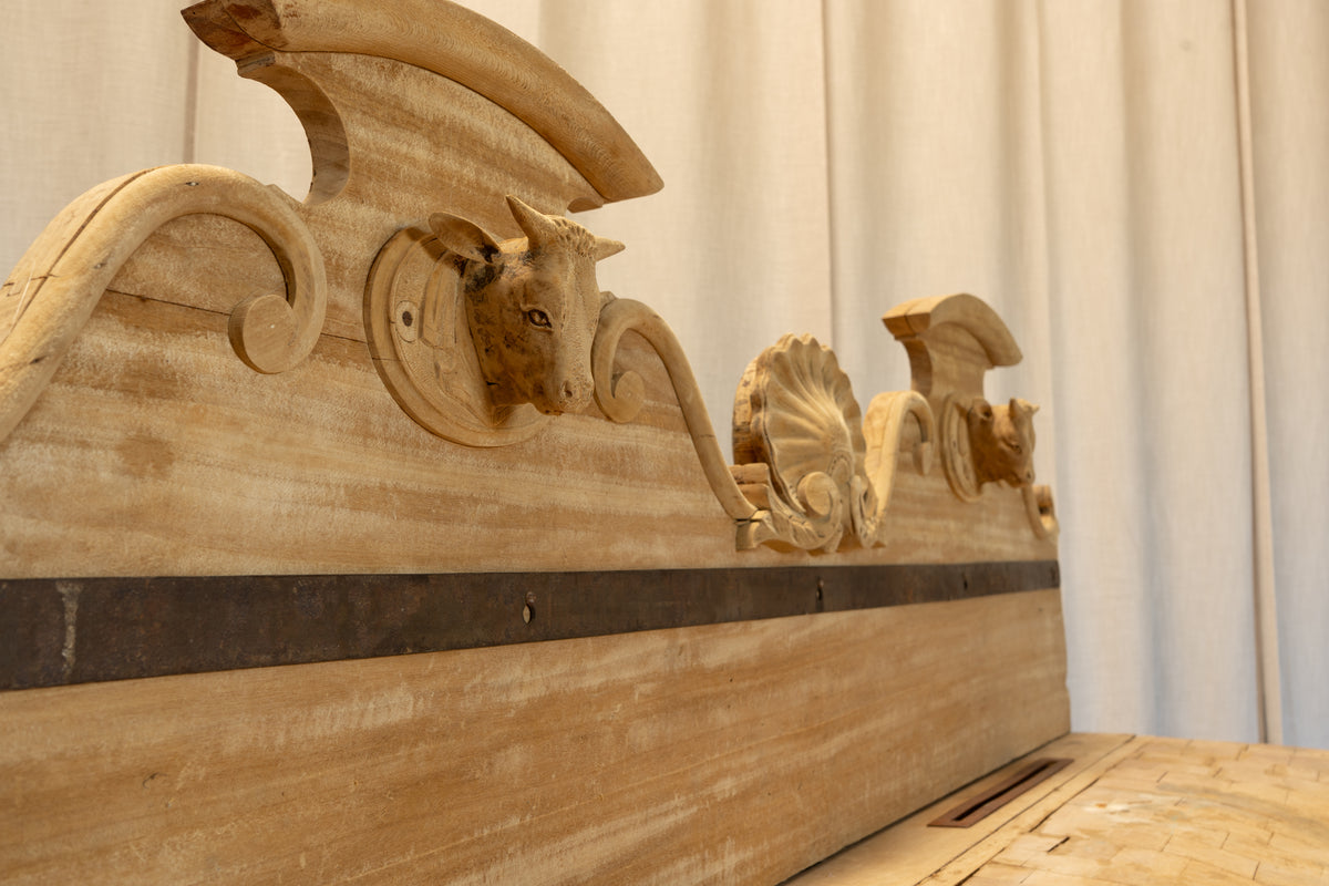 Close-up of a carved wooden headboard with ornate scrollwork, a bull head motif, and a dark horizontal inlay on a light wood panel, set against a cream curtain background.