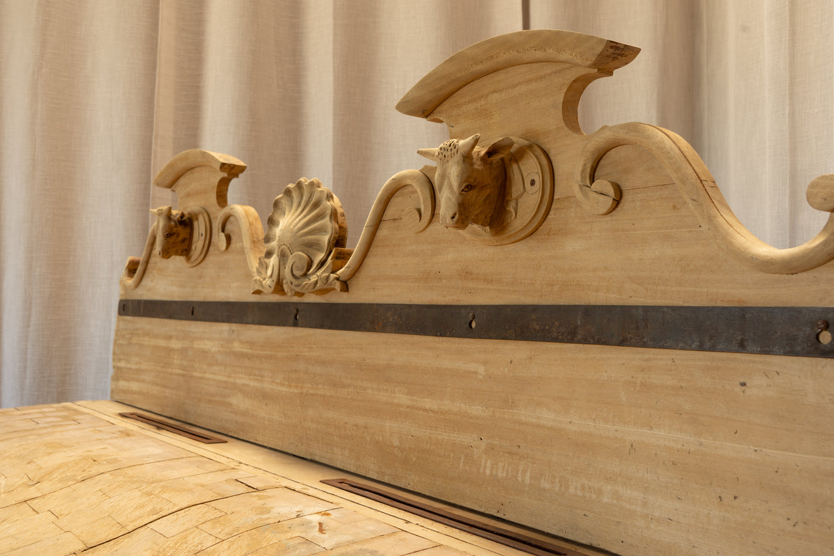 Close-up of an ornate carved wooden headboard in light natural wood with a dark horizontal metal accent strip, set against a neutral draped fabric backdrop.