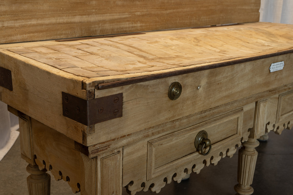 A close-up of a rustic wooden chest or bench with carved details, featuring two drawers with brass handles and dark metal corner brackets on a light wood surface.