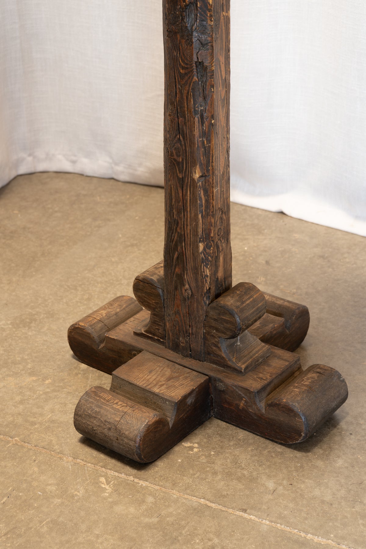 A close-up of a dark brown wooden stand or pedestal base with a thick central post and four rounded support feet, photographed indoors against a light fabric backdrop and concrete floor.