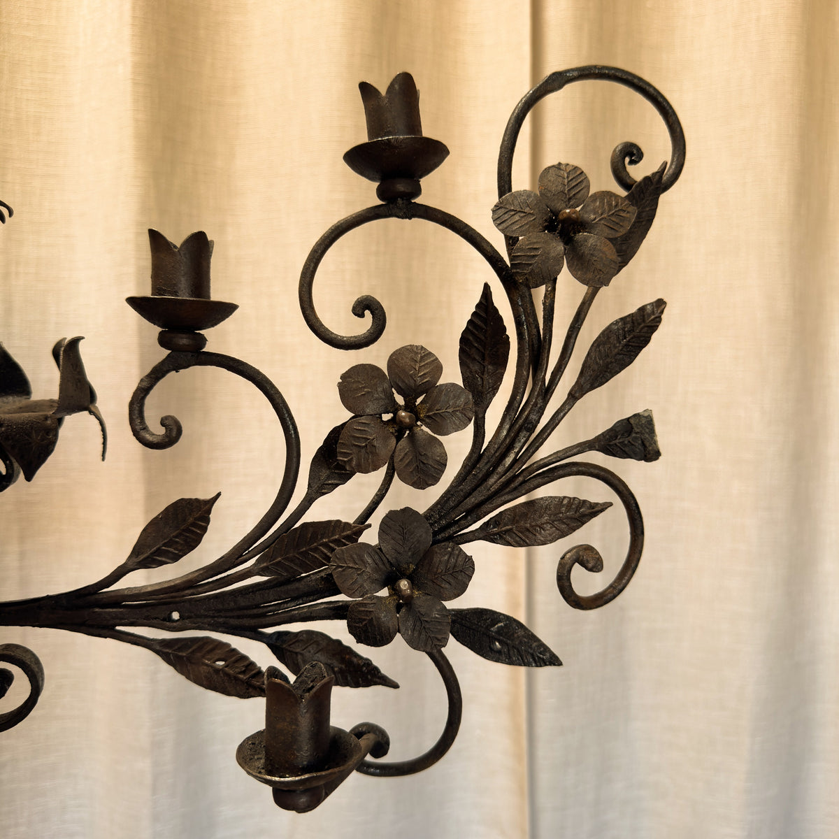 A close-up of a dark metal candelabra branch with sculpted floral and leaf details, set against a soft beige curtain background.