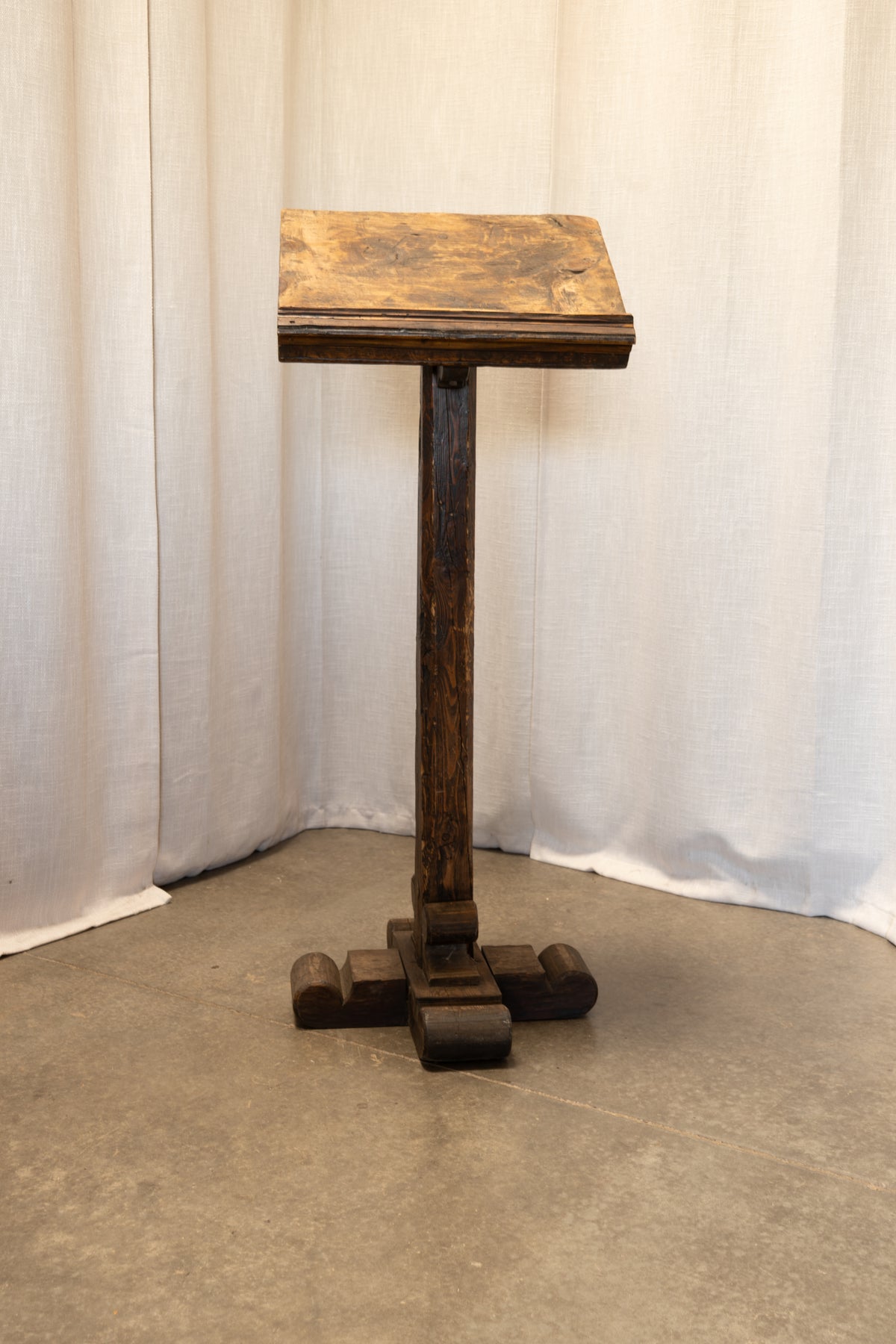 A dark brown wooden lectern stand with a square top, turned legs, and a cross-shaped base, photographed against a draped white fabric backdrop on a concrete floor.