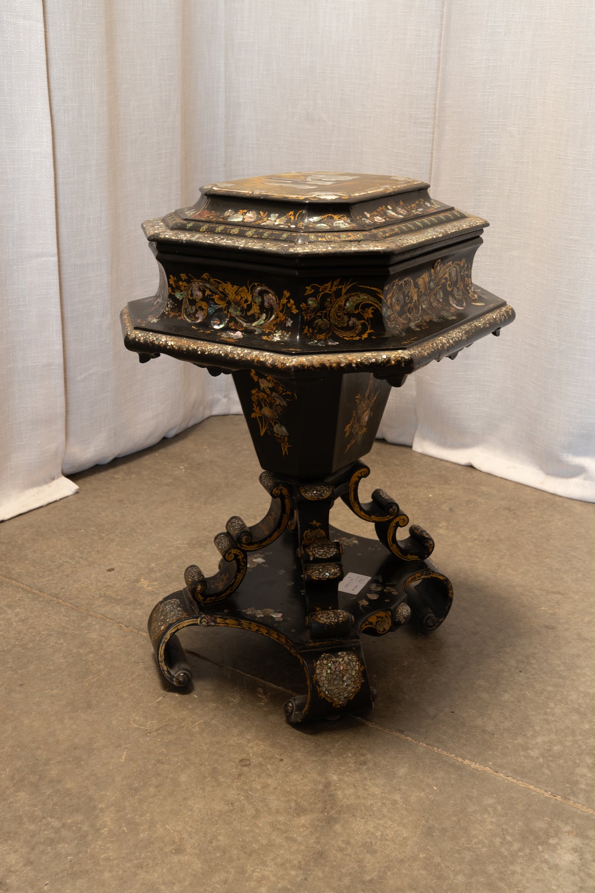 An ornate black and gold octagonal pedestal table with carved floral detailing and a tiered base, photographed against a white curtain backdrop on a concrete floor.