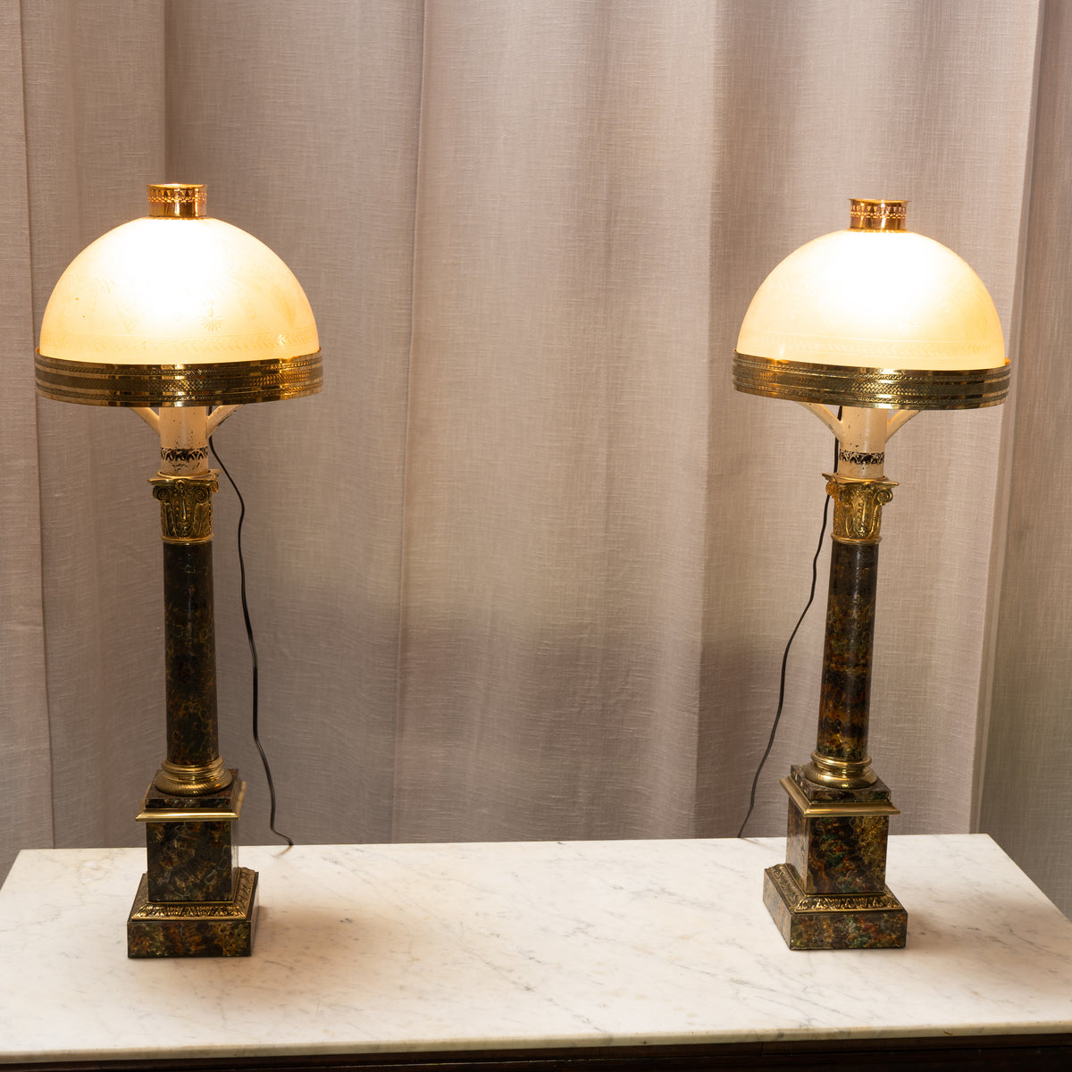 Two matching table lamps with oval cream glass shades, dark marble columns, and brass accents, displayed side by side on a white marble surface against a beige curtain backdrop.