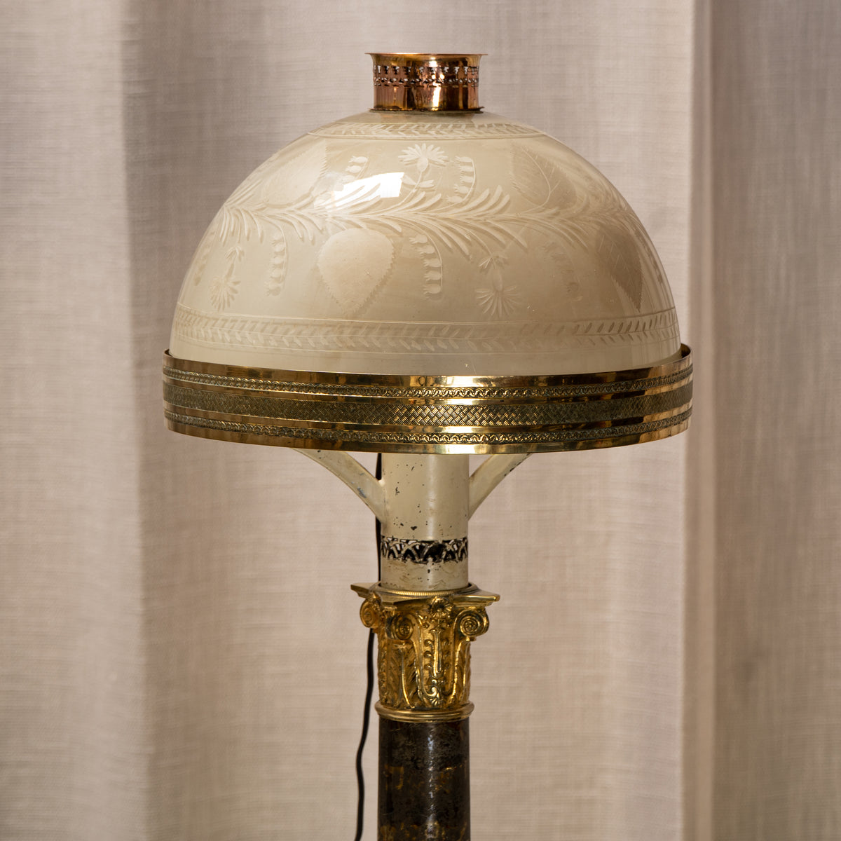 An ornate vintage-style table lamp featuring a cream-colored glass dome shade with etched floral motifs, a wide gold-toned metal band, and a dark column base with gold decorative accents, set against a neutral beige curtain background.