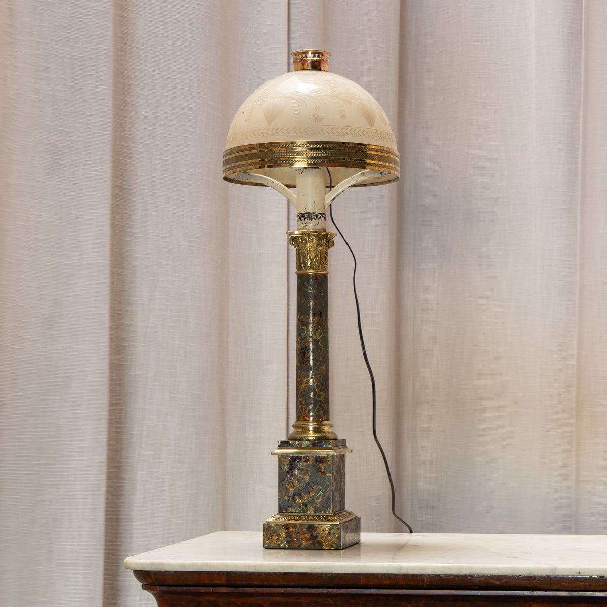 An ornate table lamp with a cream-colored glass dome shade, gold-toned metal trim, and a dark marble column with gold leaf detailing, set on a white marble-topped surface against a beige curtain background.