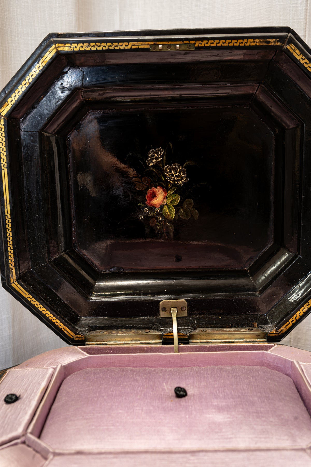 An antique octagonal music box or decorative box shown open, with a glossy black lid featuring a small floral painting in the center and gold trim, resting on a pink velvet-covered base.
