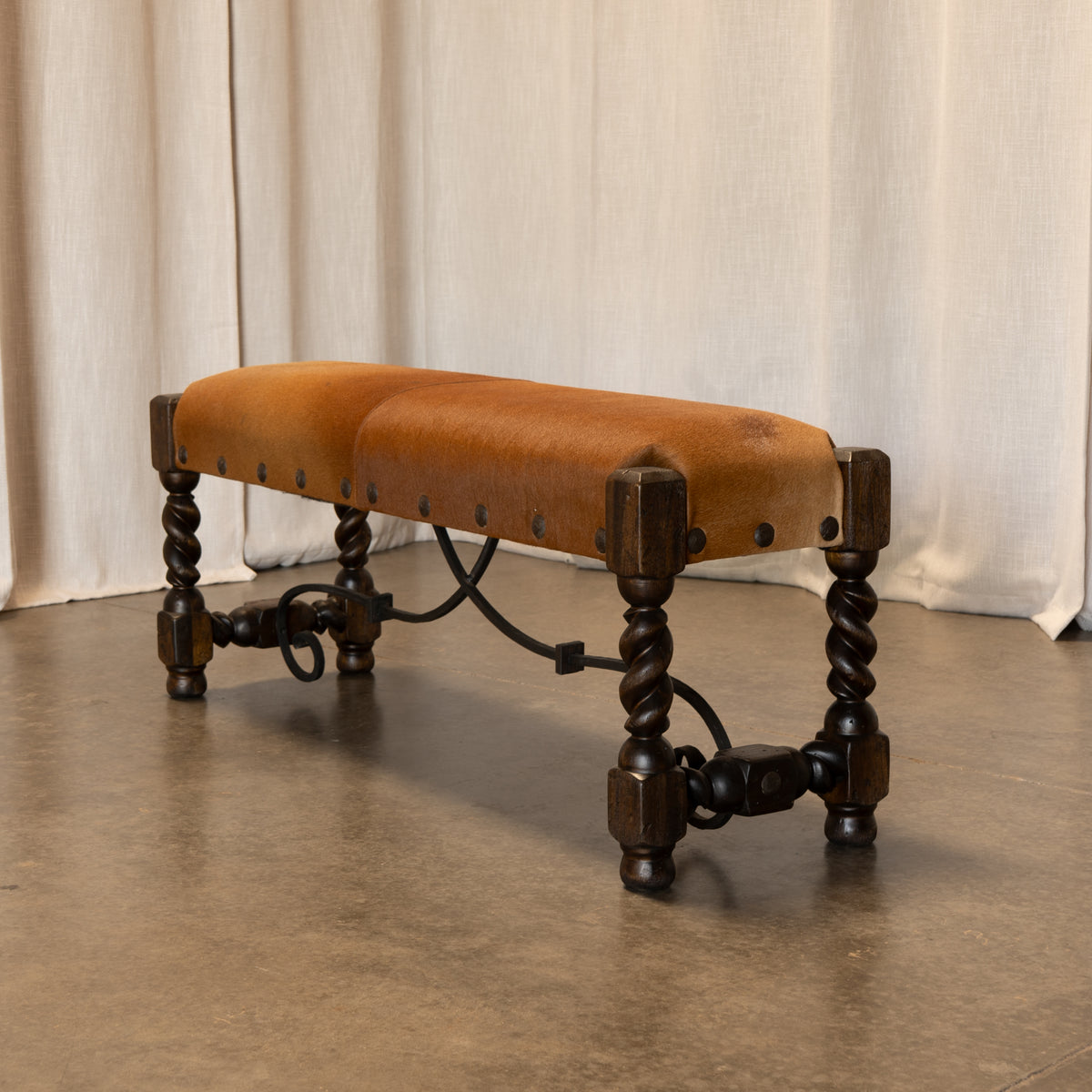 A rustic wooden bench with a tan cowhide upholstered seat, dark carved legs, and metal hardware, set against a neutral draped curtain backdrop.